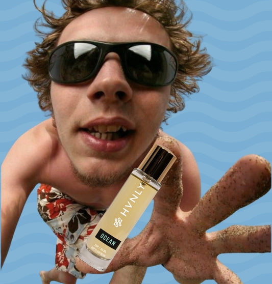 Person in swim trunks and sunglasses holding the bottle labeled Hvnly Ocean against a blue sky.