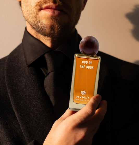 Man holding a perfume bottle labeled 'Oud of the Gods' by Hvnly against a neutral background showcasing the sleek design and the size of the bottle