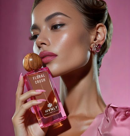 Woman holding a pink 'FLORAL CRUSH' perfume bottle by HVNLY with a wooden cap against a pink background
